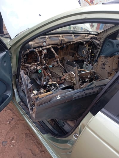 The image shows the interior of a disassembled car, possibly a sedan, with its dashboard removed and various electrical components, wiring harnesses, and structural elements exposed. The car's exterior is a light green color, and the doors are open. The scene appears to be outdoors, with reddish-brown dirt ground visible around the vehicle. The car's hood is also open, revealing more of the engine compartment. The overall condition of the interior suggests it is undergoing repair or has been dismantled. The image is taken from the passenger side of the car, looking towards the driver's side. There is no indication of people actively working on the car in this frame. The location is identified as Kontagora, Nigeria, suggesting an urban or semi-urban setting. The image is a high-angle shot, looking down into the car's interior.
