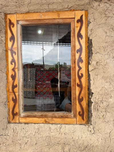The image shows a close-up of a window set into a rough, textured wall, likely made of mud or adobe. The wooden window frame is simple and appears weathered, with decorative brown swirling patterns painted onto the vertical pieces of the frame. The windowpane reflects the sky, clouds, and some of the interior of the room. A white, torn piece of fabric hangs loosely inside the window.
Reflected in the window, a person is visible, holding a smartphone to take a selfie. They are wearing a dark top and denim shorts. Inside the room, glimpses of patterned fabric and a red lattice structure can be seen.
The wall surrounding the window is uneven and cracked, suggesting an older structure or a traditional building method. The overall impression is of a rustic setting. Based on the context of Кызыл-Туу, Kyrgyzstan, this window could be part of a traditional dwelling, possibly a yurt, or a more permanent structure built with local materials. The natural and somewhat unrefined appearance of the wall and window frame hints at a rural or less modernized environment.