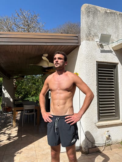 This image depicts an outdoor residential scene in Hofit, Israel, on a clear, sunny day. An adult male, appearing to be in his 20s or early 30s, stands shirtless with a muscular physique and short dark hair, wearing dark athletic shorts with a white drawstring and a "Nike Trail" logo on the left leg. He is positioned on light-colored tiled paving, with his hands on his hips, looking slightly upward and to his left.
To the left of the male subject, a covered patio area is visible, featuring a dark brown slatted wooden ceiling with a white ceiling fan. Further back within the patio, a wooden dining table is surrounded by various chairs, and a potted plant is on the ground. Beyond the patio, lush green foliage and trees are present, with string lights strung within the greenery. Strong sunlight casts distinct shadows onto the tiled ground in this area.
To the right of the male subject, a white, textured stucco wall of a building is prominent. Mounted on this wall are a white rectangular floodlight and, closer to the ground, an electrical outlet with cables connecting to a dual-port adapter. A window with dark horizontal louvered blinds is also set into the wall. Above the patio roof and the building, a clear blue sky is visible, against which the bare, leafless branches of several trees are silhouetted.
