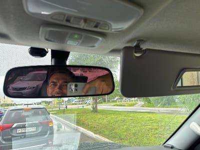 The image is taken from the interior of a car, looking forward through the windshield and a rearview mirror. The driver, a man with a smile, is visible in the rearview mirror, holding a smartphone to take the photo. He is looking directly at the camera. A purple car is reflected in the rearview mirror as well.

The setting appears to be a street in a town, possibly Usinsk, Russia, given the context. Cars are lined up on the road, suggesting traffic. The weather seems overcast, and the ground appears wet, possibly from recent rain. Green grass and trees are visible on the side of the road, indicating an outdoor, possibly suburban or park-like area. There are some buildings and streetlights in the background.

On the rear windshield of the car in front of the viewer, a license plate is visible with the text "P 318 BY 11 RUS". The overall impression is a casual snapshot taken by a driver during a traffic stop or pause. The man's expression is friendly and relaxed.