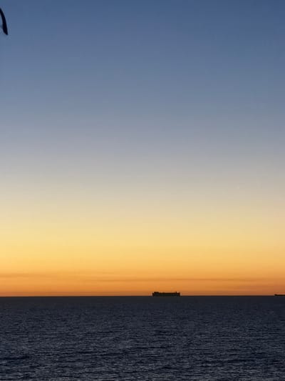 The image displays an ocean scene at City Beach, Australia, during twilight. The sky, occupying the upper portion of the frame, features a gradient transitioning from a pale blue at the zenith to a vibrant orange-yellow at the horizon line. A singular, bright white point of light is visible in the upper-mid right section of the orange-yellow sky. The lower half of the image consists of dark blue ocean water, appearing calm with minimal surface undulations. Two cargo vessels are present as dark silhouettes on the flat horizon: a prominent vessel is centered in the frame, and a second, smaller vessel is partially visible on the far right edge. A dark, indistinct, curved object is partially visible in the extreme upper left corner of the frame, against the blue sky. No individuals are visible.
