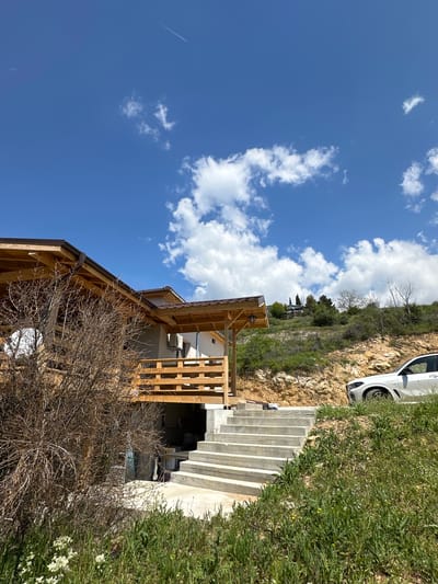 The image depicts a construction site in Lyubovka, Bulgaria. A partially built wooden structure stands prominently, possibly a house or an extension. To the right, concrete stairs lead up a grassy slope. A white car is parked near the top of the slope. The sky is blue with scattered white clouds. A bare tree in the foreground partially obscures the wooden structure.