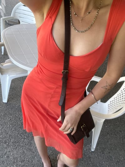 A view from directly above shows a fair-skinned person sitting on a white plastic chair with their legs to the left and torso angled toward the viewer. The person is wearing an orange dress, a brown leather belt, and a layered necklace with beads and a small pendant. They are holding a small brown purse in their left hand, which has a large ring on one of the fingers. The person has a tattoo of a starfish on their left forearm. The background consists of gray pavement and other white plastic chairs. The scene appears to be outdoors. The location is not discernible as being in Kelowna, Canada, or any other specific location.