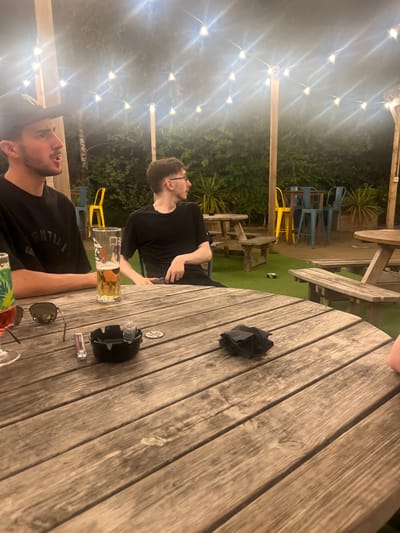 Two men sit at a wooden table under string lights. One man holds a beer, while the other looks off into the distance. An ashtray, sunglasses, lighter, and a crumpled napkin also sit on the table. A few tables and chairs are visible in the background, surrounded by plants and shrubbery.