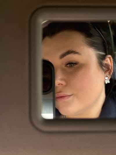 A person's face is reflected in a rectangular mirror with rounded corners. The person has fair skin, dark hair, and is wearing earrings. The environment appears to be indoors, with a neutral-colored frame surrounding the mirror. The image provides no information to identify a location in the city of Yugo-Kamskiy, Russia.