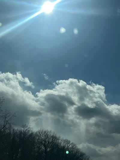 The image depicts a daytime scene in the Town of Fishkill, United States of America. The sky is partially clear blue on the left, with the sun shining brightly, producing a strong glare and light rays. Two translucent, circular artifacts are visible near the sun, possibly lens flare or dust particles. The right side of the image is dominated by a large, puffy, gray-white cloud formation. Bare tree branches are visible in the lower right corner, silhouetted against the clouds. A circular green artifact is present near the edge of the tree canopy. The overall impression is one of a sunny day with clouds gathering.