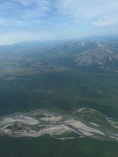 This aerial image depicts a vast, undeveloped landscape with a winding river system dominating the foreground. The river is braided, with numerous sandbars and islands covered in vegetation. Dense, dark green forests cover the banks of the river and extend into the middle ground. In the distance, rolling hills and mountains can be seen, some with patches of snow on their peaks. The sky is a pale blue with scattered white clouds. The image appears to be a natural, remote area, possibly in a region like Siberia or the Russian Far East, given the mention of Splavnaya, Russia, a city known for its river port and industrial activity. However, the depicted landscape does not show any urban development or human structures that would be indicative of Splavnaya itself, suggesting it is a view of the natural environment surrounding such a city rather than the city proper.