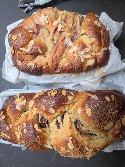 Two loaves of braided bread sit on pieces of white parchment paper, on a dark grey countertop. The bread is golden brown and topped with sliced almonds and coarse sugar. The loaf on the left appears to have a red filling, possibly jam. The other loaf has a dark filling. A grey folded cloth is visible on the edge of the scene, to the left of the leftmost bread. Location: Eeklo, Belgium, is not possible to determine from this image.