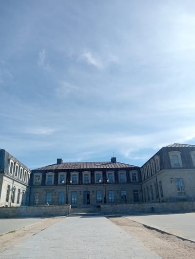 The image displays the Palacio de los Duques de Alba in Piedrahíta, Spain, under a clear blue sky with sparse, wispy white clouds. The grand, U-shaped stone building occupies the midground, its central section and two wings extending towards the viewer. The façade is constructed of light grey stone, featuring numerous rectangular windows with light-colored frames, some equipped with dark iron railings. The central entrance is accessed by a series of stone steps. The building is topped with dark, steeply pitched mansard roofs, likely of slate or metal, punctuated by multiple dormer windows and at least two visible chimneys. Two ornate black lampposts are positioned directly in front of the building's central section. The foreground consists of a wide paved area with light grey rectangular paving stones forming a path leading directly to the palace steps. This path is flanked by sections of bare, sandy earth. A low stone wall acts as a barrier separating the paved area from the building's base. No individuals or vehicles are present in the scene.