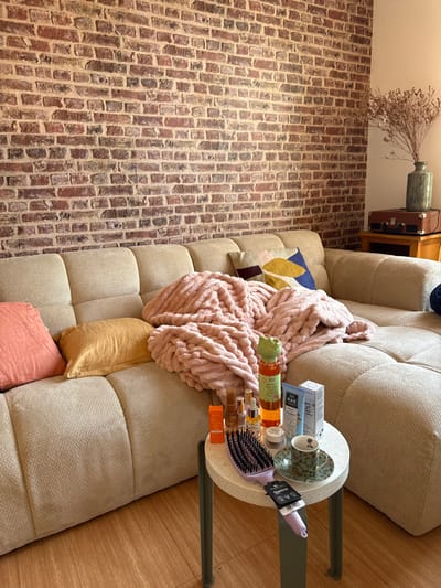 The image depicts an interior residential scene in Saint-Cyr-l'École, France. The primary subject is a large, modular, light-beige sectional sofa occupying the lower and middle left sections of the frame. A thick, ribbed, light-pink blanket is draped loosely over a central section of the sofa. Two solid-colored throw pillows are visible on the sofa to the left: one coral-pink and one golden-yellow. Further to the right on the sofa, a patterned throw pillow with geometric shapes in blue, yellow, and burgundy is visible.

In front of the sofa, slightly to the right, a round, white marble-patterned side table with three dark-green metal legs holds an array of personal care items. These include several bottles of liquids, a white box labeled "MAE," a purple hairbrush with a black hang tag, and a green and white patterned teacup and saucer. A green bottle with a frog-shaped cap is prominently placed among the items.

The background features a exposed brick wall with varying shades of red, brown, and gray bricks and light mortar, covering the majority of the upper frame. In the far upper right corner, a small, light-brown wooden cabinet is visible. On top of this cabinet rests a dark-brown portable record player, and behind it, a muted green ceramic vase holds a dried arrangement of light-brown plant material. The floor is light-colored wood. No individuals are present in the scene.