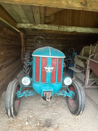 In a rustic wooden shed in Sielachowskie, Poland, a turquoise and red vintage tractor is parked. The tractor's front grill is a prominent red with vertical slats, and it features a circular emblem with a winged symbol. Two round headlights are mounted on the front axle. The tractor has large, treaded tires on the rear and smaller ones on the front, with red rims visible on the front wheels. The shed's interior is constructed from rough-hewn wooden logs and planks. Behind the tractor, other agricultural machinery is visible, including a fan and what appears to be a threshing machine. The ground is covered with dirt and scattered debris. No people are present in the image.