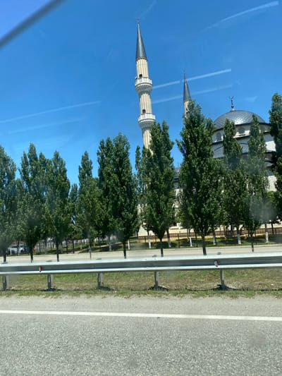 The image captures a daytime view of a mosque, likely in the city of Koshkel'dy, Russia, as suggested by the presence of minarets and a dome characteristic of Islamic architecture. The mosque features light-colored stone walls and a dark, tiered roof. Two prominent minarets, one taller than the other, rise into a clear blue sky. The shorter minaret is topped with a crescent moon symbol. A row of tall, slender green trees stands in front of the mosque, partially obscuring its lower levels. A black metal fence with white posts runs along the property line behind the trees. In the foreground, a grey asphalt road is visible, with a white line marking a lane. A metal guardrail separates the road from a grassy area. The scene appears to be viewed from a moving vehicle, as suggested by the slight reflections and haziness on the surface, possibly a window. No people are visible in the frame.
