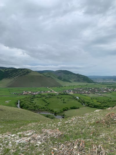 This is a vertical, high-angle, eye-level, full shot of a landscape in Khurai-Nur, Russia, on a cloudy day. In the foreground, the rocky, grassy slope of a hill is visible, with patches of white and yellow wildflowers. A winding river flows through a lush green valley in the middle ground. On the banks of the river are scattered trees and shrubs. Beyond the river, a village with houses and a road is nestled at the foot of rolling green hills. The hills are covered with trees in some areas and grasslands in others. In the background, more rolling hills recede into the distance, with a hazy sky filled with gray clouds. There are no people or animals visible in the image.