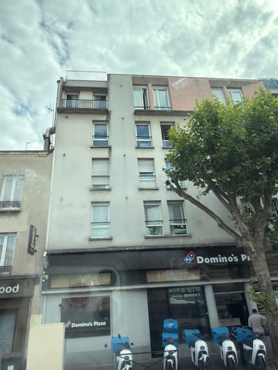 This image captures a street view of a building with a Domino's Pizza restaurant on the ground floor in Alfortville, France. The building is light-colored and has multiple stories with numerous windows. A large tree with green foliage is prominently situated to the right of the building, partially obscuring it. The Domino's Pizza storefront features its distinctive logo and branding, with a slogan that appears to be in French. In front of the restaurant, several white electric scooters are parked, some equipped with blue delivery boxes bearing the Domino's logo. A person in a light-colored shirt and dark pants is seen walking away from the scooters towards the right side of the frame. The sky is partly cloudy.