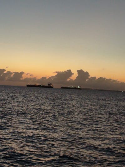 Two cargo ships are sailing on the ocean at dusk, near the city of Gulhi Falhu, Maldives. The sky is a gradient of light blue and orange, with scattered clouds. The water is dark and choppy, reflecting the dim light of the sky and the ships' lights. The ships are long and narrow, with lights illuminating their decks and superstructures. The scene is tranquil and serene, with a sense of calm and solitude.