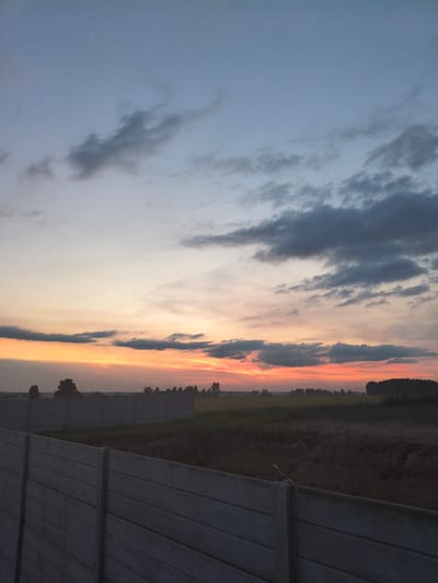 This photograph captures a sunset over a rural landscape near Galonki, Poland. The sky is a gradient of colors, transitioning from a soft blue at the top to hues of orange and red near the horizon. Wispy clouds are scattered across the sky, some appearing dark gray and others catching the fading sunlight. In the foreground, a concrete barrier, likely a fence or wall, stretches across the lower portion of the image. Beyond the barrier, the land is a mixture of fields and distant trees. The overall scene is tranquil and evokes the end of the day.