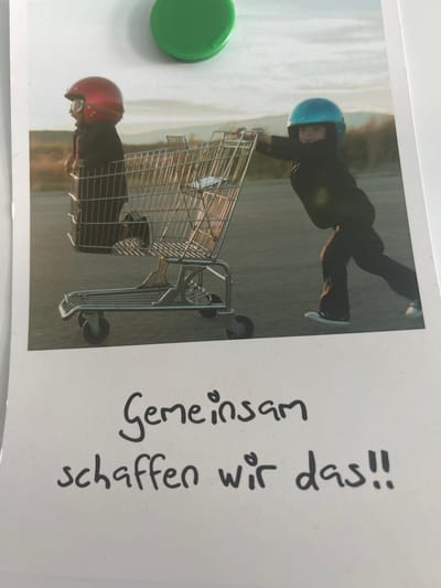 Two young children, one wearing a red helmet and goggles and the other a blue helmet, are playing with a shopping cart outdoors. The child in the red helmet is inside the cart, and the child in the blue helmet is pushing it. They appear to be on a paved surface, possibly a street or parking lot, with a blurred background of trees and hills. The image is formatted as a Polaroid photo, and below the picture, there is handwritten text in German that reads "Gemeinsam schaffen wir das!!", which translates to "Together we can do it!!". There is also a green circular object, likely a magnet, at the top of the image. The location in Glanegg, Austria, is not specified within the image itself.