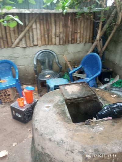 The image depicts an outdoor scene in Dokan Tofa, Nigeria. The foreground features a concrete structure, possibly a wellhead, with a metal cover. Adjacent to this, a black rectangular object, possibly a storage box, is visible, topped with two orange plastic containers, one larger and one smaller, and a blue lid. To the left, a blue plastic chair is positioned on a brown decorative block. Further back, a black plastic chair is placed in front of a light blue small table. Another blue plastic chair is situated to the right, near a tree trunk. The background consists of a concrete wall with a bamboo-like fence above it. Green foliage is visible at the top of the image. A date stamp "06/16/2026 15:56" is present in the lower right corner.