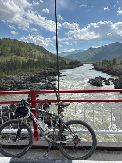 A bicycle is resting against a red and gray metal railing, situated on a bridge above a river. The river has a churning, white appearance. In the background, a landscape of green-covered mountains and a cloudy sky can be seen. The bicycle is gray with a water bottle attached to its frame, and a white helmet is strapped to the handlebars. A thick black cable stretches across the top of the frame. The location is the city of Edigan, Russia.