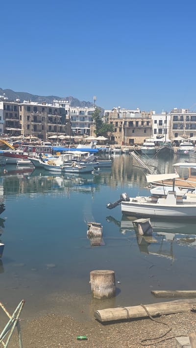 The image depicts a harbor scene in Kyrenia, Northern Cyprus. Numerous boats of various sizes are moored in the calm, blue water. Many of the boats are white with blue accents. In the background, there are several multi-story buildings with stone and white facades, characteristic of Mediterranean architecture. Some buildings have balconies and awnings. Mountains rise in the distance behind the buildings, under a clear blue sky. The foreground shows a rocky shore with a wooden log and some metal structures. The water reflects the sky and the boats.