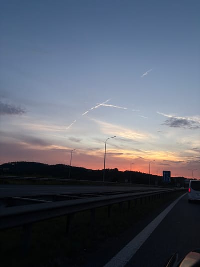The image depicts a sunset view from a highway in Mirošovice, Czechia. The sky is a gradient of soft blues and pinks, with faint clouds and two contrails from airplanes forming a cross shape. A dark, silhouetted tree line runs along the horizon, with a few streetlights visible along the highway. A guardrail separates the lanes of the road. A white van is visible in the right lane, and a blue traffic sign with arrows indicating multiple lanes is also present. The foreground shows a dark asphalt road with a white lane marker.