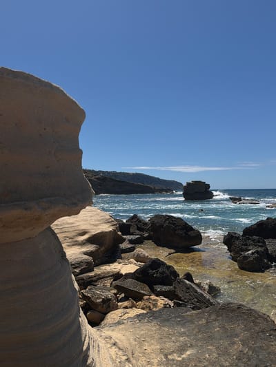 The image depicts a rocky coastline in Llucmajor, Spain. In the foreground are light-colored, eroded sandstone rocks. In the background, the sea is visible with dark-colored rocks scattered along the shoreline. A rock formation rises from the sea. A person is swimming in the water. The sky is blue with a few clouds. The photo is taken in a rotated, portrait orientation.