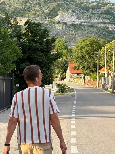 A man, facing away from the camera, walks along a paved road in Rajićevići, Montenegro. He wears a short-sleeved shirt with vertical brown stripes on a white background and khaki cargo shorts. His right hand is visible, holding a dark object, and a black watch is on his left wrist. He wears thin-rimmed glasses. The road has a white dashed line marking its center and shoulder. To the left of the road, a dark metal gate and some greenery are visible. On the right side of the road, there are stone houses with red tile roofs, stone walls, and lush green trees. The background features a steep, rocky mountainside with sparse vegetation and power lines overhead. The scene is illuminated by natural sunlight, suggesting it is daytime.