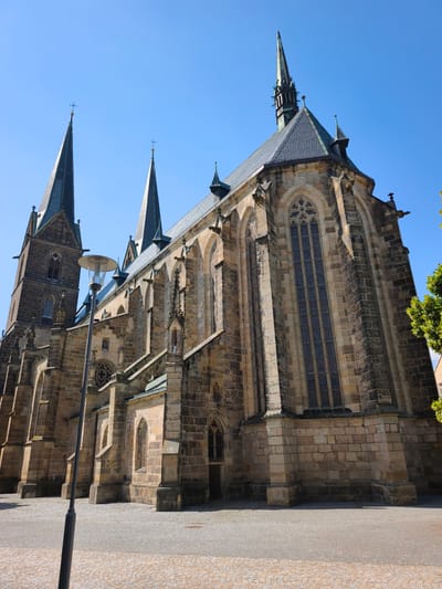 A low-angle, wide shot captures the exterior of a grand Gothic-style church under a clear, bright blue sky, suggesting a sunny day. The church is constructed from rough, tan stone, with multiple pointed spires adorned with crosses. The architecture features elaborate stonework, including arched windows with stained glass, gargoyles, and flying buttresses. A tall lamppost stands in the foreground, to the left of the church, its simple design contrasting with the ornate building. The ground in front of the church is paved with cobblestones.
The scene is serene and static, with no people visible. The sunlight casts distinct shadows, indicating it is likely midday or early afternoon. The architectural style and the clear, sunny weather contribute to a sense of historical grandeur and peacefulness. The church appears to be a significant landmark in the town of Vysoké Mýto, Czechia. No text is visible in the image.
