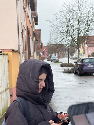 An outdoor scene in the city of Meistratzheim, France, depicts a young female in the foreground on a wet, overcast day. The subject, viewed in profile from the right, occupies the left half of the frame. She wears a black, hooded puffer jacket with the hood fully up, obscuring most of her dark hair. Her visible right hand holds and interacts with a smartphone; her left hand is also engaged with the device. A dark backpack strap is visible over her left shoulder.
To the subject's immediate left, a light yellow-orange building wall with white trim and a white wooden gate is visible. The background reveals a residential street, characterized by wet asphalt, indicating rain or recent precipitation. A dark purple Renault Zoé (license plate GD-040-GG) is parked on the right side of the street, facing away from the viewer. Further down the street, a silver hatchback is parked on the left, facing towards the viewer, with additional vehicles visible in the distance.
The street is lined with multi-story buildings featuring pitched roofs and varied pastel facades, including light peach, orange-brown, yellow, and pink, consistent with traditional Alsatian architecture. These buildings have windows, some with closed shutters. A prominent leafless deciduous tree stands on the right side of the street, adjacent to a dark, ornate lamppost. Several small, low-lying shrubs are planted in circular beds along the roadside. The sky is uniformly grey and overcast.