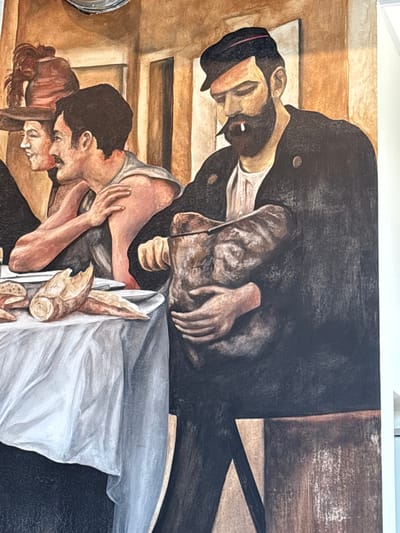 This is a close-up, angled photograph of a mural depicting several figures in a somber, intimate scene. The mural appears to be painted in a realistic style with muted, earthy tones.
The central focus is on two couples. In the foreground, a man and woman are embracing closely, their faces touching in what seems to be a tender or sorrowful moment. The woman has reddish-brown hair pulled back, and the man has dark hair. They are partially covered by a white cloth, suggesting a scene of rest or grief. Next to them, a loaf of bread is prominently featured, broken into pieces.
Above and behind them, another couple is depicted. The man, wearing a dark coat and a hat, leans over the woman, whose head is bowed. Their hands are intertwined or touching, and the overall impression is one of deep emotion, possibly loss or farewell. The background consists of abstract, block-like shapes in shades of brown and beige, resembling steps or architecture.
The lighting in the mural is dramatic, with strong shadows and highlights that add to the emotional weight of the scene. The overall mood is serious and contemplative. There is no visible text in the image. The location context provided is Elsenfeld, Germany, which could suggest the mural is part of a public artwork or historical commemoration.