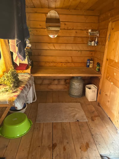 The image depicts the interior of a wooden room, likely a Russian banya or sauna, located in Чуй-Атасево, Russia. The walls and ceiling are constructed from horizontal wooden logs. A wooden bench runs along the back wall, above which is a narrow shelf holding several bottles of toiletries and a small wire rack with additional items. A large oval mirror is mounted on the log wall above the shelf.
To the left of the bench, a table is covered with a patterned tablecloth. On the table sits a bunch of dried leafy branches, possibly for use in a sauna. Behind the table, several folded towels and what appears to be a small appliance are visible. A yellow curtain hangs to the left of the table.
On the wooden floor, a light brown, rectangular mat lies in the center of the room. To the left of the mat, a large, bright green plastic basin is placed on the floor. Beneath the bench, a large, galvanized metal milk can stands next to a white cardboard box. The wooden floor shows some water stains and appears worn. A wooden door with a simple design is visible on the right side of the image. The overall impression is of a functional, rustic bathing space.