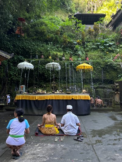The image depicts a spiritual site in Bangli, Indonesia, likely Tirta Empul Temple or a similar water temple. Three individuals are visible in the foreground, facing away from the camera, engaged in prayer or meditation. One person, on the left, kneels with hands clasped, wearing a light blue and white shirt and a patterned skirt with a green sash. In the center, a woman in a patterned sarong and yellow sash meditates cross-legged on a mat. To her right, a man in a white shirt and white cap sits cross-legged on a mat.
Behind them is a raised altar draped in black fabric with a yellow, pleated valance. Several decorative white and yellow umbrellas are positioned above the altar, from which water streams down, creating a cascade effect. Small figures, possibly religious statues, are arranged on a ledge above the waterfalls. The background is a lush, green, densely vegetated cliff face, suggesting a natural grotto or ravine. Additional figures are visible further back, near what appears to be a bathing pool with a fence. The ground in the foreground is wet and speckled with flower petals. The overall atmosphere is serene and contemplative.