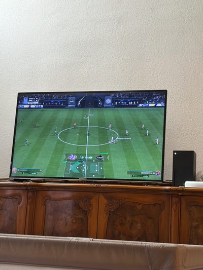 This image, captured in Eschau, France, depicts an indoor domestic scene. A large flat-screen television occupies the center, displaying a soccer video game in progress. The game scoreboard indicates "KOR 0 - 0 GRE" at "00:00". Virtual players, identifiable by green and white/red kits, are positioned on a striped green digital pitch. On-screen overlays show player names such as "10 LEUCHTER" and "BEERENSTEYN 10", alongside a tactical map in the lower center. To the right of the television, a black Xbox Series X console stands upright, adjacent to a smaller white electronic device. The television rests on a dark brown wooden credenza, which features multiple ornate carved panels across its front. The background consists of a light-colored, textured wall. The upper portion of a light brown, tufted sofa is visible in the immediate foreground.