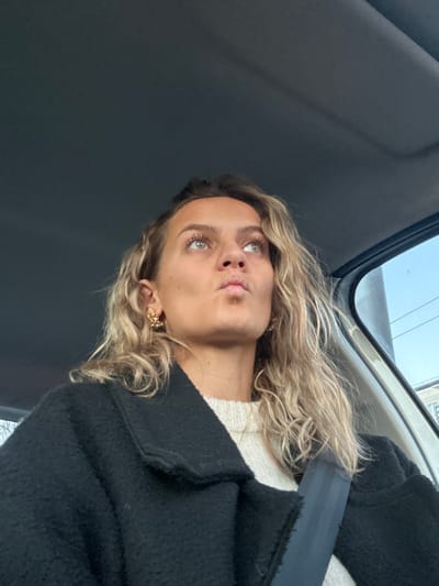 A woman with wavy blonde hair sits in a car, wearing a white sweater and a black coat. A seatbelt crosses her chest. She gazes upwards with a slight pout, revealing striking green eyes. Delicate gold earrings adorn her ears. The car interior is visible, with a window showing a glimpse of the outside.