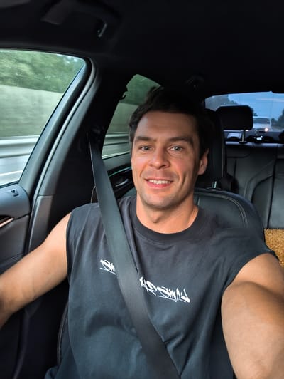 A man is seated in the driver's seat of a car, smiling at the camera. He is wearing a black t-shirt with a white graphic design on the front. The car's interior is visible, including the black leather seats and a seatbelt. Outside the car window, trees and a road can be seen, suggesting the car is in motion. The lighting indicates it is daytime. The image does not contain any information that would allow for the determination of its location in Vasilevo, Russia.