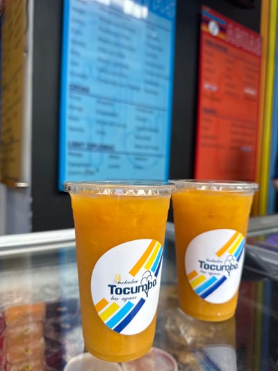 The image shows two cups of orange juice on a glass counter. Both cups have the logo "Helados Tocumbo las aguas," and are located in front of a menu board with a blue background and the word "AGUA" in white, and a second menu board with an orange background and the word "ALMUERZOS" in white. The location is at the coordinates (latitude: 25.657, longitude: -100.439) in Ciudad Santa Catarina, Mexico.
