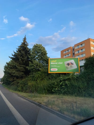 The image captures a billboard advertising "Dr. Max" in Strakonice, Czechia. The billboard, featuring a pair of sunglasses, is placed alongside a road, with lush green trees and a multi-story orange building in the background. The sky is a vibrant blue with scattered white clouds. The road, marked with a white line, suggests a setting on the outskirts of the city.