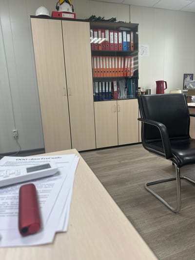 The image captures an indoor office environment, featuring furniture and organizational items, suggesting an administrative or storage area. The perspective is from a desk looking towards a cabinet and chair. The image appears to be rotated 90 degrees clockwise from its natural upright orientation.
In the foreground, a light-colored wooden desk is visible, holding a stack of white papers, a white calculator, and a red marker or pen. Visible text on the top paper includes "ООО «БашТехСнаб»" (OOO BashTechSnab).
Towards the midground, a black office chair with shiny chrome legs is positioned. Behind the chair and extending across the background is a large, light-colored wooden cabinet. The upper part of the cabinet has glass doors, revealing rows of brightly colored ring binders in red, orange, and blue. Some binders have labels, with "Входной контроль 2026" (Incoming control 2026) and "ПЛУНЖЕРЫ" (Plungers) clearly legible on different binders. The lower section of the cabinet has solid wooden doors.
To the left of the cabinet, on the light grey wood-look laminate floor, a white hard hat with yellow earmuffs is placed, next to a red binder. Some dark green artificial foliage is visible behind the cabinet. On the white, horizontally paneled wall above the cabinet, a framed document or award hangs, displaying the word "ГРАМОТА" (Gramota). A red pitcher or thermos is also partially visible on top of the cabinet behind the chair. A white electrical outlet with a cord is present on the wall near the floor.
No individuals are present, and no specific activity is taking place, giving the scene a quiet and unoccupied feel. The visible Russian text on documents and the award is consistent with the provided location context of Gubkinsky, Russia. The time of day and weather are not discernible from the indoor setting.