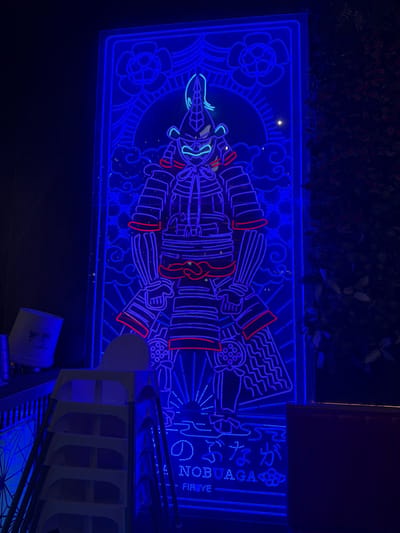 The image shows a large, brightly lit blue neon sign against a dark background. The sign depicts a stylized, almost robotic-looking figure in traditional Japanese attire, possibly a samurai or a character from a play. The figure is outlined in blue neon, with some accents in red. It is set within a decorative frame that includes floral elements and swirling patterns, reminiscent of traditional Japanese art.
To the right of the figure, there is text in neon. Part of it appears to be Japanese characters, and below that, in English, "NOBUAGA" is clearly visible, followed by "FIR Eye". The overall impression is that of an advertisement or a decorative element for a Japanese restaurant or entertainment venue.
The setting appears to be outdoors at night, given the darkness and the illumination provided by the neon sign. The dark foliage in the upper portion of the image suggests it might be located in an area with greenery, perhaps a courtyard or a street. The sign is large and commands attention. Below the sign, a section of a modern building or structure with curved white elements and metal framework is visible, indicating an urban or commercial environment. There are also some cylindrical objects in the foreground, one of which appears to be a trash receptacle. The bright blue light cast by the sign creates a vibrant and somewhat dramatic atmosphere. The location context provided is Carretera De San Martín, Spain, which is a street name.
