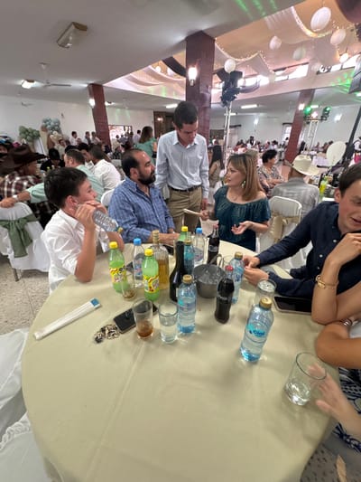 A slightly high-angle, medium indoor shot depicts a group of people seated around a table at an event or gathering. The location is identified as Chipilo de Francisco Javier Mina (Chipilo), Mexico. The people are diverse, with varying ages and appearances. The individuals at the table are dressed in smart casual attire.
The table is covered with a light-colored tablecloth. It is laden with bottles of water and beverages. There are also glasses and personal items like phones and jewelry scattered across the table.
In the background, more people are seated at other tables in a large room. Decorations such as white drapes, lights, and balloons adorn the space. The overall impression is that of a celebratory occasion.