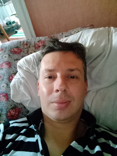The image is a selfie of a man lying on a floral patterned sofa with a white blanket. He is wearing a black and white striped shirt. The background appears to be a room with a window, and a wall with an orange-brown textured surface. The man's face is partially visible, with his eyes looking towards the camera. There are no discernible elements in the image that would indicate a specific location in the city of Hamarnia, Ukraine.