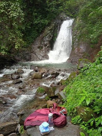 The image shows a waterfall in Tawangmangu, Indonesia, at the coordinates -7.655 latitude and 111.135 longitude. The waterfall cascades down a rocky cliff face, creating a pool of water at its base. The water flows through a riverbed with many rocks. Lush green foliage surrounds the waterfall. A red jacket, a backpack, a blue water bottle, and a blue container are placed on the rocks in the foreground. The water is clear and flowing rapidly. There are no people visible in the image. The scene suggests a peaceful and serene natural environment.