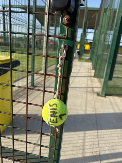 The image shows a close-up of a green metal fence with rusty bars. Hanging from the fence by a rope and a key ring is a tennis ball. The tennis ball is lime green with black writing on it, which appears to read "TENIS." The background is out of focus, but it appears to be an outdoor sports facility, possibly a tennis or paddle tennis court, with green fencing and a paved walkway. The sky is clear and blue. The overall lighting suggests it is daytime, likely late afternoon given the long shadows cast by the fence. No people are visible in the image. The scene is likely located in Vidreres, Spain, as indicated by the prompt, suggesting this is a public or private recreational area within that municipality.