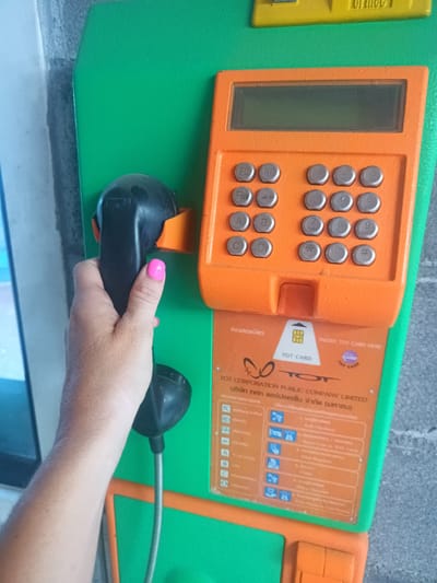 The image shows a person holding the handset of a public telephone. The phone is green and orange with a keypad and a display screen. It is labeled "TOT CARD" and includes Thai writing, indicating it is located in Thailand, specifically Surat Thani City Municipality. The woman has a pink nail polish. The phone also has instructions on how to insert a TOT card.