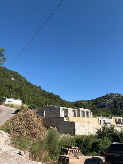 This image depicts an outdoor scene in Markovići, Montenegro, under a clear, bright blue sky, suggesting a sunny day. The perspective is a bit tilted, looking up a hillside dotted with lush green trees and rocky outcrops. In the foreground, a road winds upwards. To the right of the road, a partially constructed building is visible, made of stone and concrete, with empty window frames and balconies. The building appears to be under construction or renovation. Adjacent to the building, stacks of lumber are piled up, and some terracotta pots are scattered. Further up the hillside, more of the building's structure is visible, seemingly built into the slope. A dark-colored SUV is parked on the side of the road near the building. A small, white, shed-like structure with a single window is situated further up the road. A thin power line stretches diagonally across the sky. The overall impression is one of a rural, mountainous area with ongoing development.