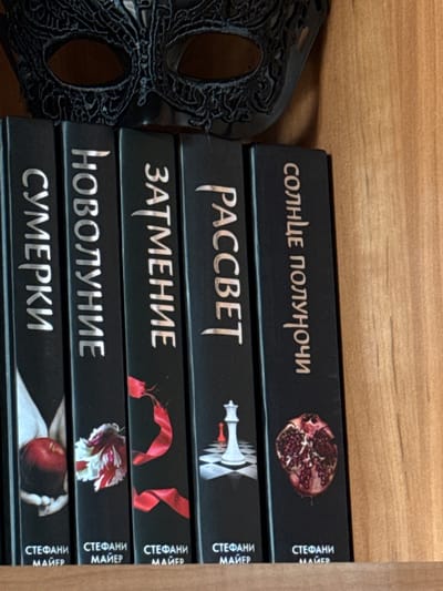The image shows a stack of five black books with silver text and illustrations on their spines. The books appear to be a series, likely novels, given the distinct titles and author names printed vertically on the right side of each spine.
The titles, in Cyrillic script, are: "СОЛНЦЕ ПОЛУНОЧИ" (Midnight Sun), "РАССВЕТ" (Breaking Dawn), "ЗАТМЕНИЕ" (Eclipse), "НОВОЛУНИЕ" (New Moon), and "СУМЕРКИ" (Twilight). Below each title, the author's name "СТЕФАНИ МАЙЕР" (Stephenie Meyer) is also visible.
Each book features a unique illustration related to its title: "СОЛНЦЕ ПОЛУНОЧИ" has a half-pomegranate, "РАССВЕТ" depicts a chess piece (a white queen and a red pawn), "ЗАТМЕНИЕ" shows a red ribbon, "НОВОЛУНИЕ" has a red and white flower, and "СУМЕРКИ" features a hand holding a red apple.
To the left of the books is a black, ornate mask, possibly a masquerade mask, with intricate cut-out patterns and two eye openings. The background is a light-colored wooden surface, suggesting the books are on a shelf. The overall impression is a collection of books displayed on a wooden surface, with a decorative mask beside them. The lighting suggests an indoor setting, possibly a room with ambient light. There are no people or any indication of a specific time of day or weather conditions. The text is clearly legible on the spines of the books.