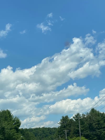 The image shows a bright blue sky filled with fluffy white clouds. A small, dark, reddish-brown smudge is visible in the upper right quadrant of the sky, possibly smoke. The lower portion of the image features a dense line of evergreen trees, and in the foreground, two wooden utility poles with wires strung between them are visible, with the tops of other poles and wires peeking through the trees. The scene appears to be taken from a roadway, as indicated by the telephone poles and wires. The image was captured in Lac-Simon, Canada.
