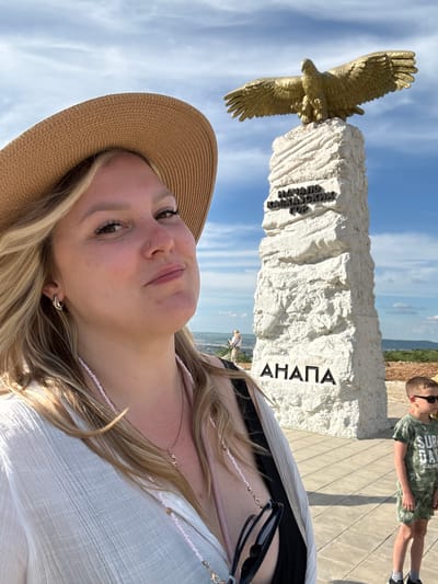 The image captures a selfie-style portrait of a woman in the foreground, with a monument and a child visible in the background. The woman, with blonde hair, wears a wide-brimmed straw hat and a white, textured top, along with a delicate beaded necklace and sunglasses hanging from her neck. She looks towards the camera with a subtle expression.
Behind her stands a significant monument composed of a large, rough-cut white stone stele topped with a golden statue of an eagle in flight. The monument features Cyrillic inscriptions. On the left side, the text "НАЧАЛО КАВКАЗСКИХ ГОР" (START OF THE CAUCASUS MOUNTAINS) is visible. On the right side of the stele, the word "АНАПА" (ANAPA) is clearly inscribed in large black letters.
To the right of the monument, a young boy in a green camouflage-patterned t-shirt, shorts, and sunglasses stands on a paved area, seemingly looking out at the distant landscape. The setting appears to be an elevated viewpoint with a clear blue sky, light cloud cover, and a hazy horizon suggesting a wide vista, possibly over a valley or sea. The sunny weather indicates it is daytime. The location cues, including "Anapa" and "Caucasus Mountains" on the monument, align with the provided context of Varvarovka, Russia.