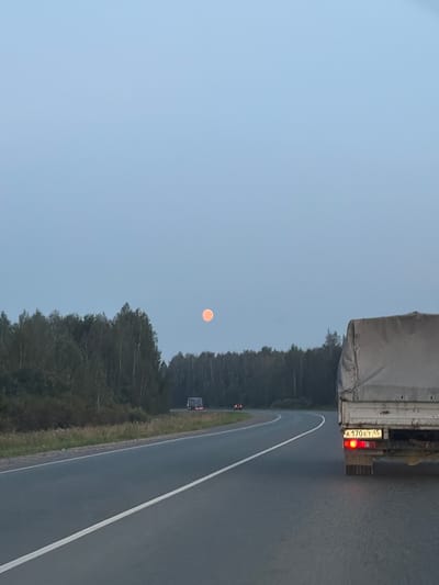 A full moon, appearing orange, is visible in the pale blue sky above a dense forest lining a curved asphalt road. The road has a white dividing line. In the foreground, the rear of a white truck is visible on the right side of the road, its license plate reading "A 170 ET 45" and its red taillights illuminated. Further down the road, a bus with illuminated lights is traveling in the opposite direction, and another vehicle with red lights is visible in the distance on the left. The environment appears to be a rural or suburban area, possibly in Shcherbakovo, Russia, given the road markings and the presence of trees and a truck. The time of day is likely dusk or early evening.