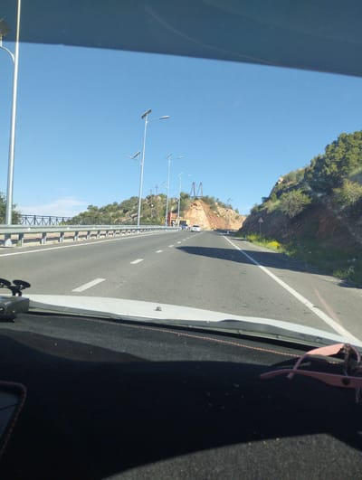 The image shows a view from inside a car driving on a highway. The highway is paved and has white lane markings. There are streetlights with solar panels lining the road. On the right side of the road, there is a steep, rocky hill covered with sparse vegetation. On the left side, there is a metal guardrail and more vegetation. The sky is clear blue. In the foreground, the dashboard of the car is visible, with a pair of pink sunglasses resting on it. There are no people visible in the image. The location is identified as Myk, Uzbekistan.